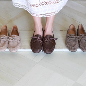 Puede incluir: Tres pares de mocasines de cuero en tonos beige claro, marr&oacute;n oscuro y topo. Cada par presenta una punta redondeada, una costura frontal fruncida y un detalle de cordones decorativo en la parte superior. Los zapatos est&aacute;n dispuestos en fila, mostrando su construcci&oacute;n de cuero suave y su dise&ntilde;o cl&aacute;sico sin cordones.