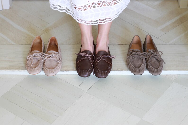 Puede incluir: Tres pares de mocasines de cuero en tonos beige claro, marr&oacute;n oscuro y topo. Cada par presenta una punta redondeada, una costura frontal fruncida y un detalle de cordones decorativo en la parte superior. Los zapatos est&aacute;n dispuestos en fila, mostrando su construcci&oacute;n de cuero suave y su dise&ntilde;o cl&aacute;sico sin cordones.