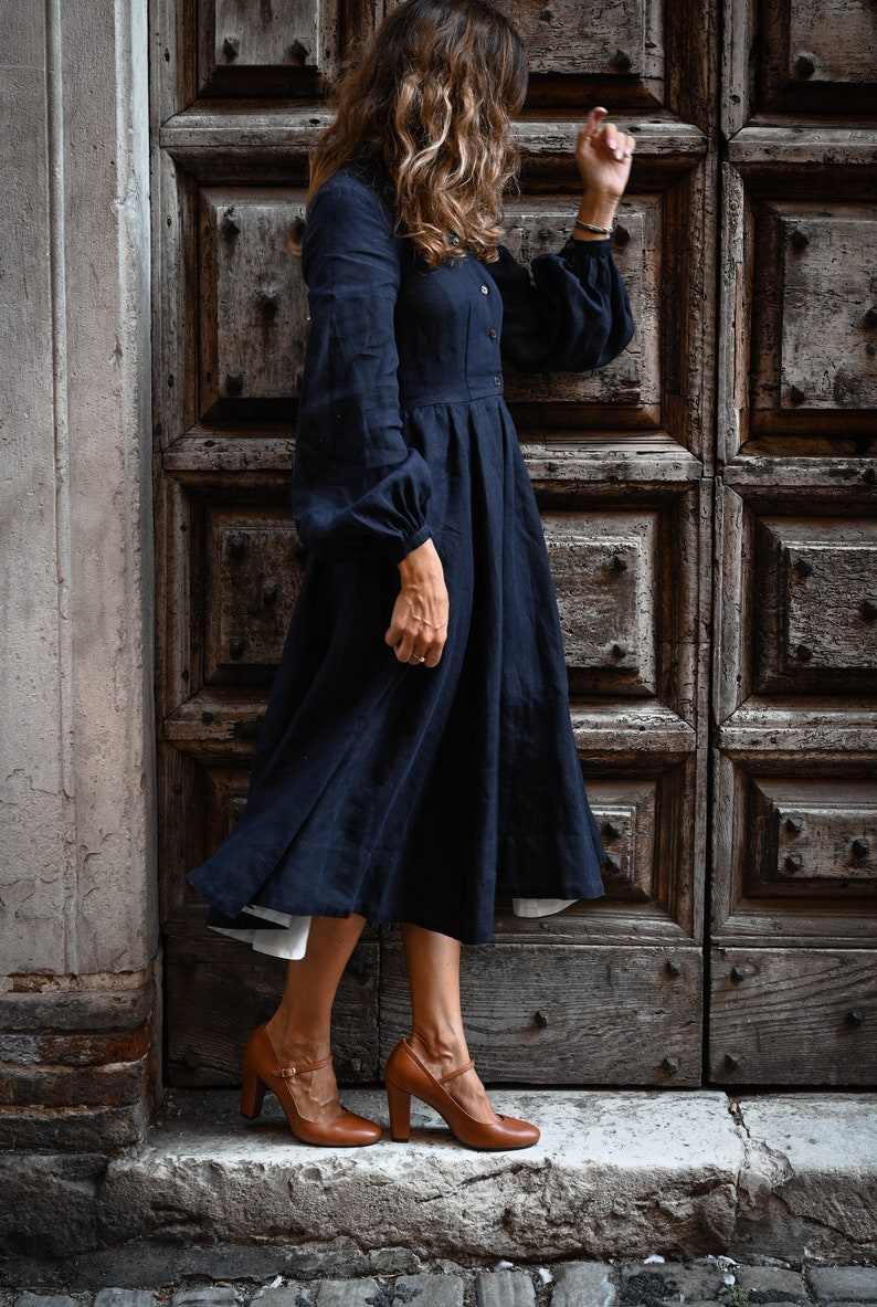 May include: A woman wearing a dark blue midi dress with long sleeves and a gathered skirt. She is wearing brown heeled shoes with ankle straps.