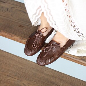 Puede incluir: Un par de mocasines de cuero marr&oacute;n oscuro con dise&ntilde;o de punta fruncida, cordones frontales con tiras finas de cuero y costuras visibles en los laterales. Los zapatos se llevan con una prenda de tela blanca con detalles de encaje.