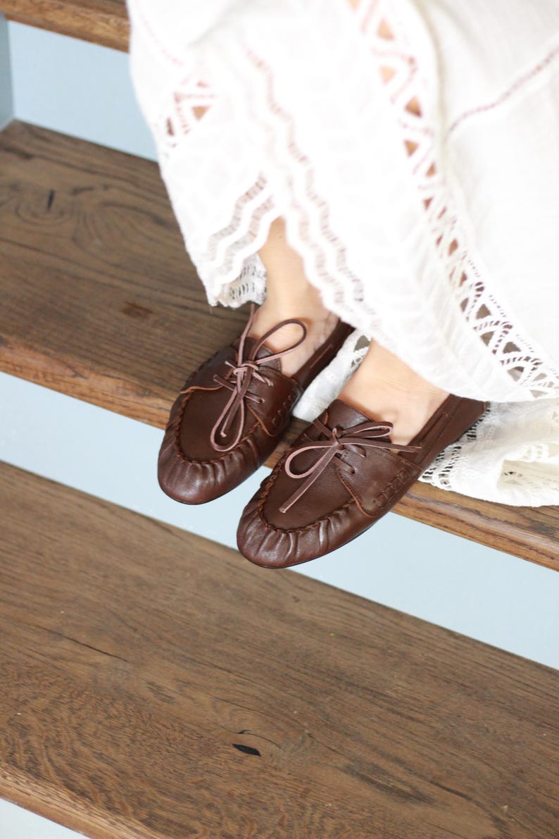 Puede incluir: Un par de mocasines de cuero marr&oacute;n oscuro con dise&ntilde;o de punta fruncida, cordones frontales con tiras finas de cuero y costuras visibles en los laterales. Los zapatos se llevan con una prenda de tela blanca con detalles de encaje.