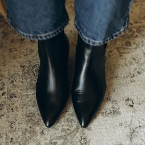 Puede incluir: Un par de botines de cuero negro con punta afilada y acabado liso, usados con pantalones vaqueros azules. Los botines presentan una silueta elegante y un dise&ntilde;o limpio, cubriendo el pie hasta el tobillo con un exterior de color negro s&oacute;lido.