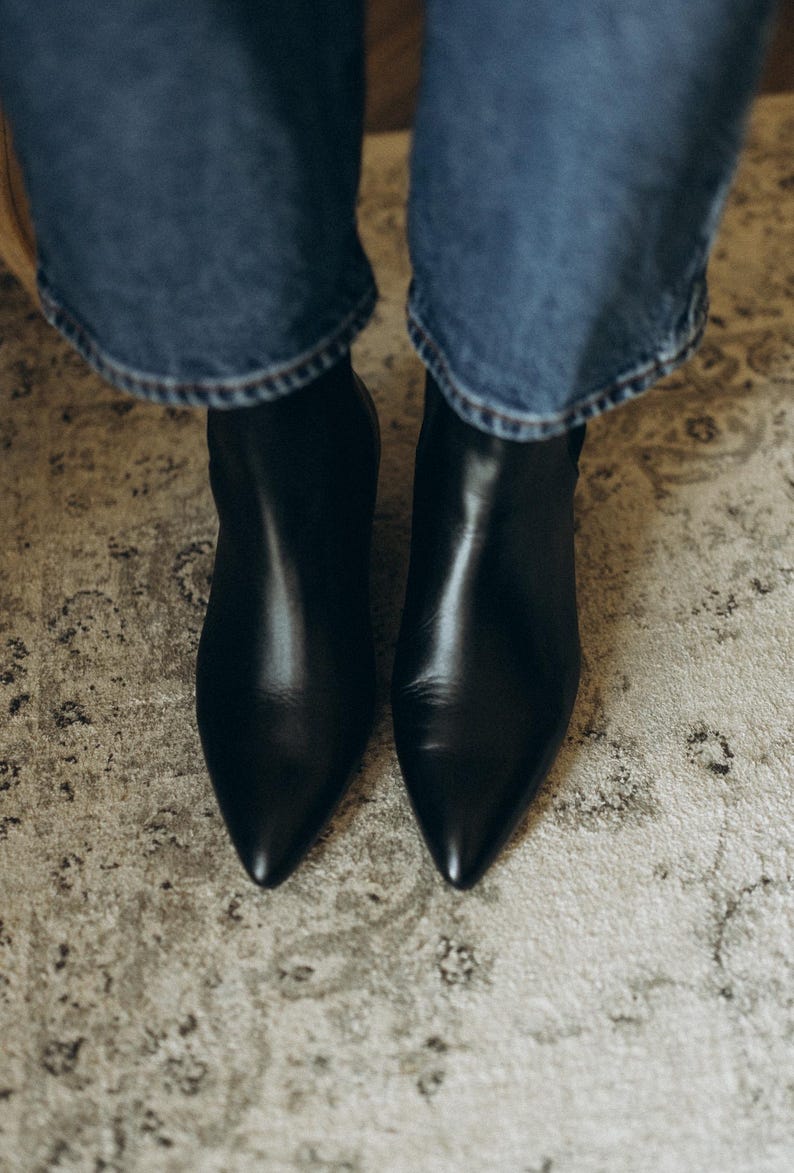 Puede incluir: Un par de botines de cuero negro con punta afilada y acabado liso, usados con pantalones vaqueros azules. Los botines presentan una silueta elegante y un dise&ntilde;o limpio, cubriendo el pie hasta el tobillo con un exterior de color negro s&oacute;lido.