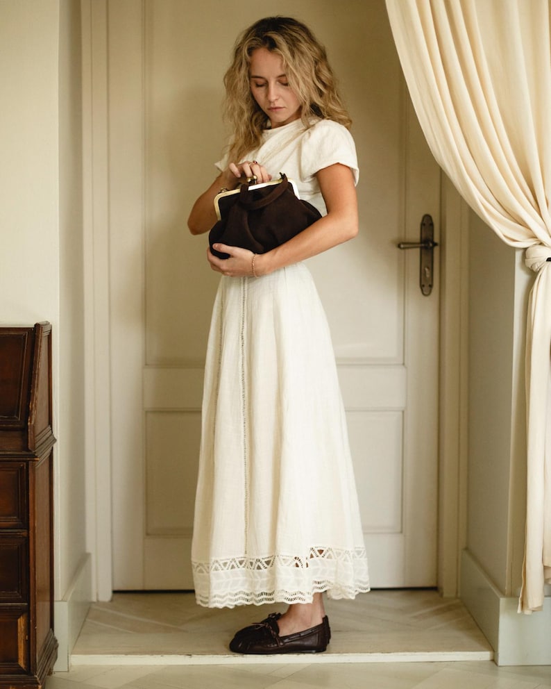 Puede incluir: Un vestido midi de color crema con mangas cortas y un dobladillo de encaje decorativo, combinado con un bolso de mano de ante marr&oacute;n oscuro con cierre de marco de metal dorado y mocasines de cuero marr&oacute;n oscuro con un lazo frontal.