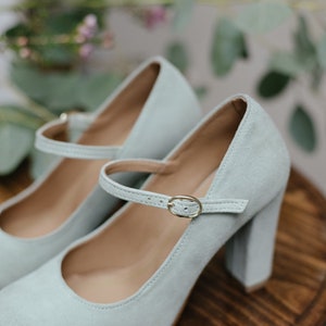 May include: A pair of light blue suede heeled shoes with a buckle strap. The shoes are on a wooden surface.