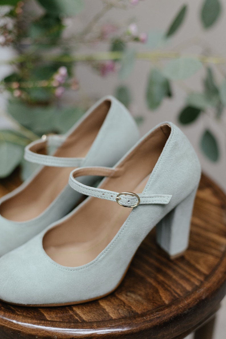May include: A pair of light blue suede heeled shoes with a buckle strap. The shoes are on a wooden surface.