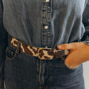 Pode incluir: Um cinto de pele de vaca castanho e branco com fivela dourada, usado sobre uma camisa de ganga e cal&ccedil;as de ganga. O cinto &eacute; um acess&oacute;rio elegante, que adiciona textura e padr&atilde;o ao conjunto.