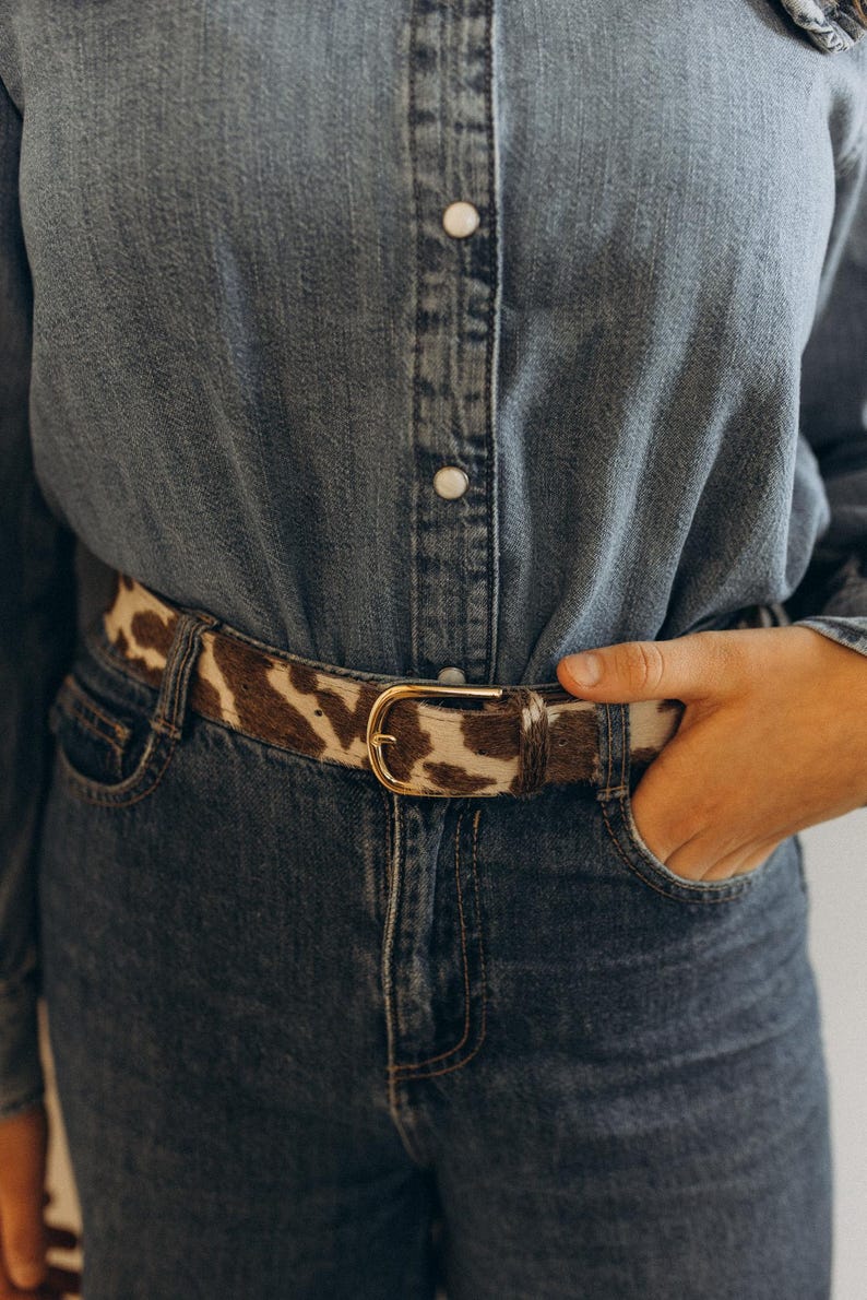 Pode incluir: Um cinto de pele de vaca castanho e branco com fivela dourada, usado sobre uma camisa de ganga e cal&ccedil;as de ganga. O cinto &eacute; um acess&oacute;rio elegante, que adiciona textura e padr&atilde;o ao conjunto.