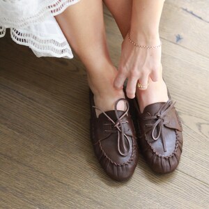 Puede incluir: Un par de zapatos estilo mocas&iacute;n de cuero marr&oacute;n oscuro con punta redondeada, una costura fruncida a lo largo del borde delantero y cordones de cuero finos atados en un lazo en la parte superior. Los zapatos tienen una estructura flexible.