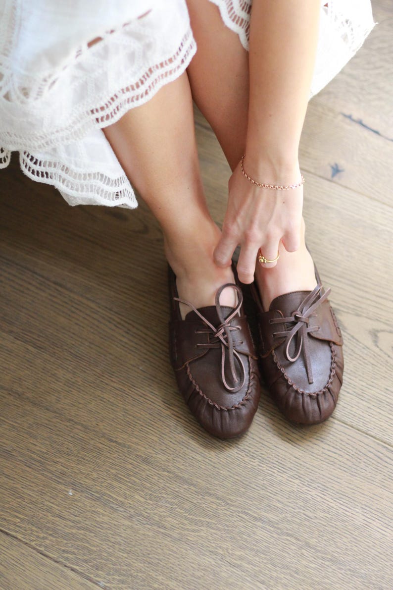Puede incluir: Un par de zapatos estilo mocas&iacute;n de cuero marr&oacute;n oscuro con punta redondeada, una costura fruncida a lo largo del borde delantero y cordones de cuero finos atados en un lazo en la parte superior. Los zapatos tienen una estructura flexible.
