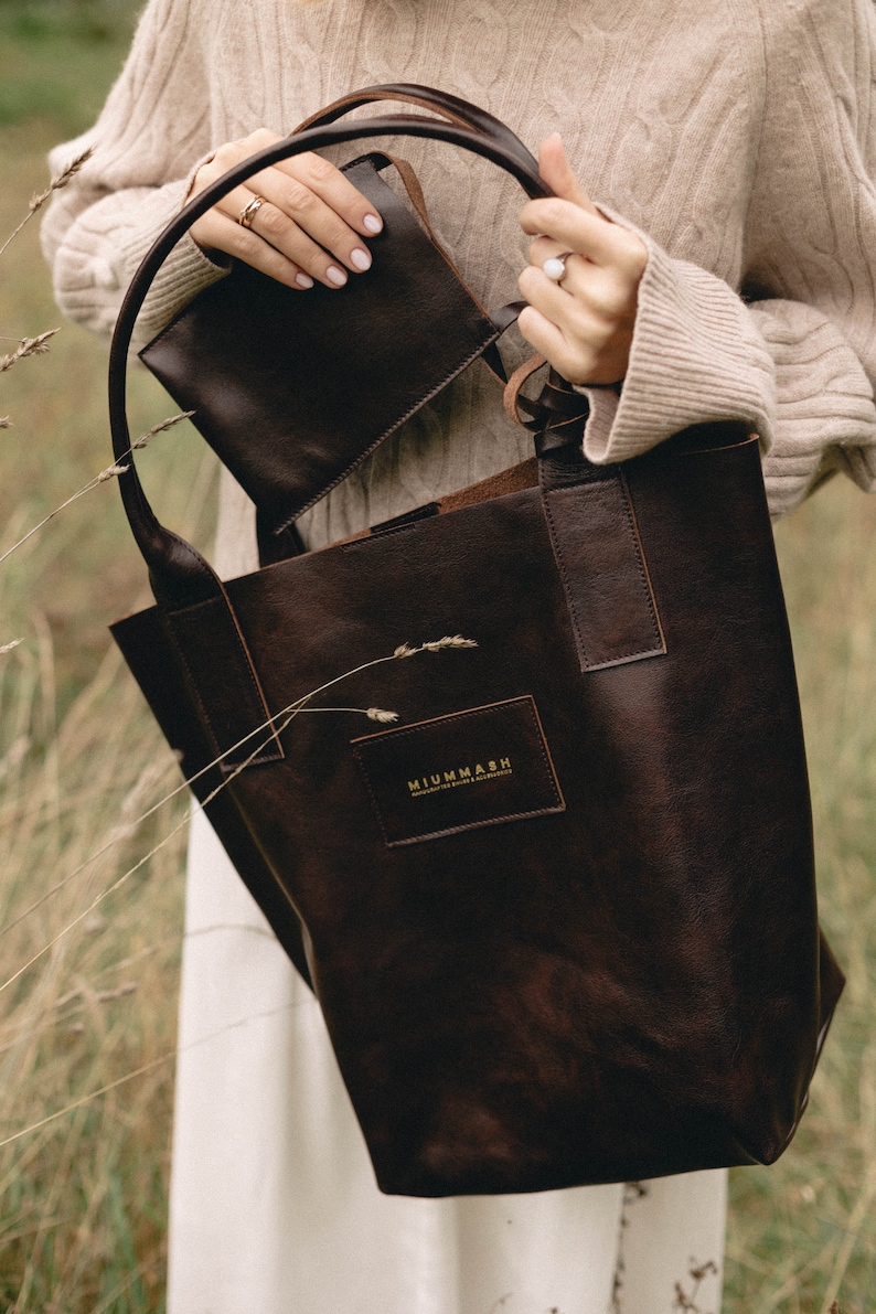 May include: A brown leather tote bag with a smaller brown leather pouch inside. The tote bag has a gold logo that reads "MIUMMASH Handmade Goods & Accessories".