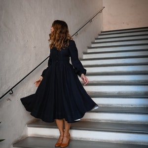 May include: A woman wearing a dark blue dress with long sleeves and a full skirt, standing on a set of white stairs. She is wearing brown shoes and has long brown hair.