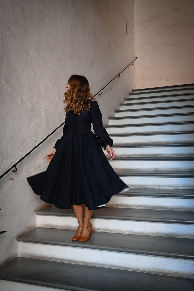 May include: A woman wearing a dark blue dress with long sleeves and a full skirt, standing on a set of white stairs. She is wearing brown shoes and has long brown hair.