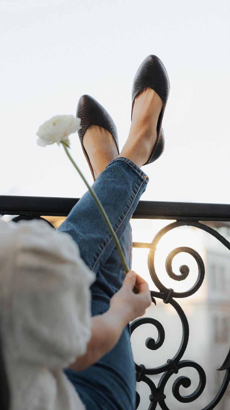 May include: Dark brown leather flats with a textured pattern. The shoes are worn with blue jeans. A white flower is held in the hand. The background includes a wrought iron railing and sunlight.