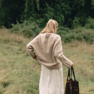 May include: A woman in a beige sweater and white skirt walks through a grassy field, carrying a brown leather tote bag.