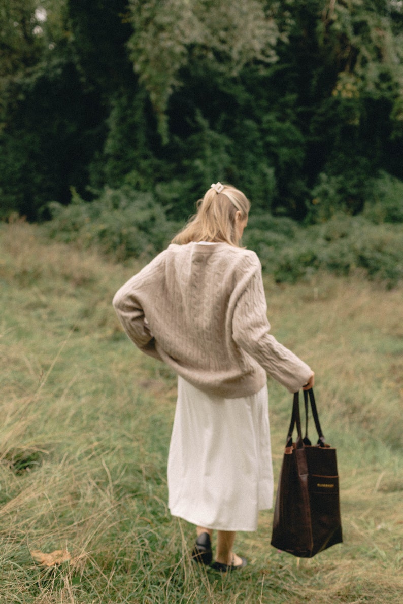 May include: A woman in a beige sweater and white skirt walks through a grassy field, carrying a brown leather tote bag.