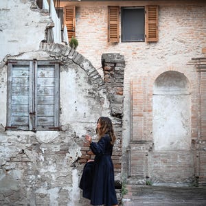 May include: A woman in a dark blue dress and brown shoes stands in front of a brick building with a weathered facade. The building has a large arched doorway and a smaller window with wooden shutters.