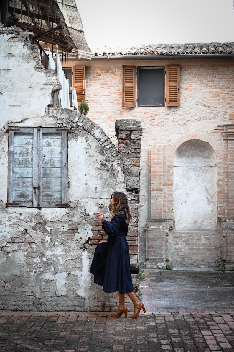 May include: A woman in a dark blue dress and brown shoes stands in front of a brick building with a weathered facade. The building has a large arched doorway and a smaller window with wooden shutters.
