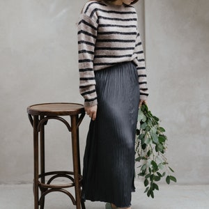 May include: A woman wearing a striped brown and black jumper, a black pleated skirt, and light blue heels. She is holding a bouquet of green leaves and standing next to a wooden stool.