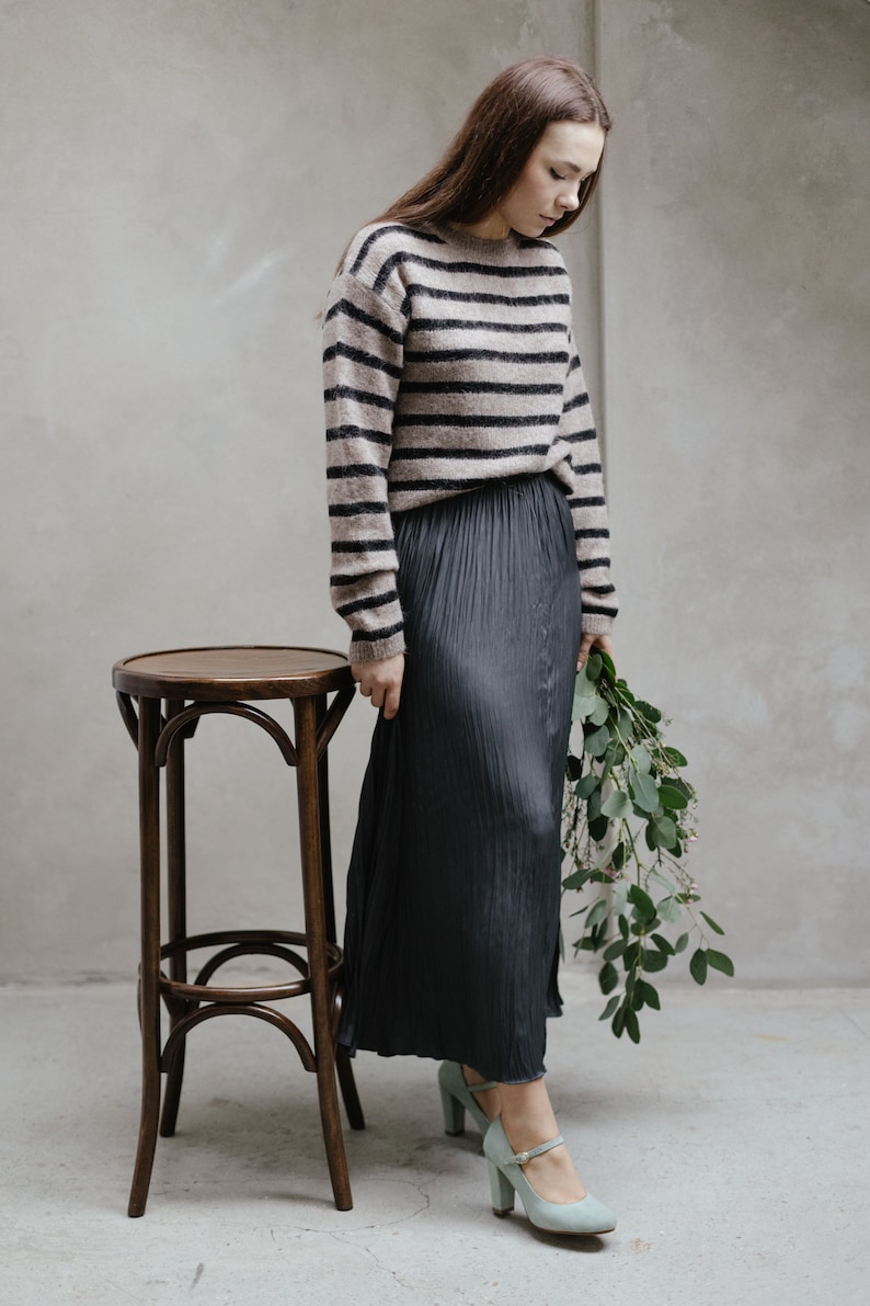 May include: A woman wearing a striped brown and black jumper, a black pleated skirt, and light blue heels. She is holding a bouquet of green leaves and standing next to a wooden stool.