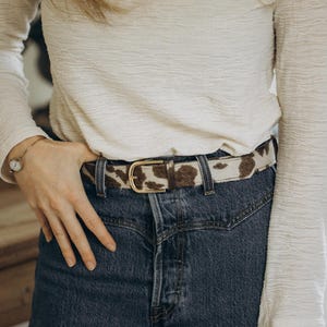 Pode incluir: Um cinto com estampa de pele de vaca e fivela dourada &eacute; usado com jeans azuis e uma camisa branca de manga comprida. O cinto apresenta um padr&atilde;o de manchas marrons e brancas. A fivela tem um design cl&aacute;ssico e arredondado.