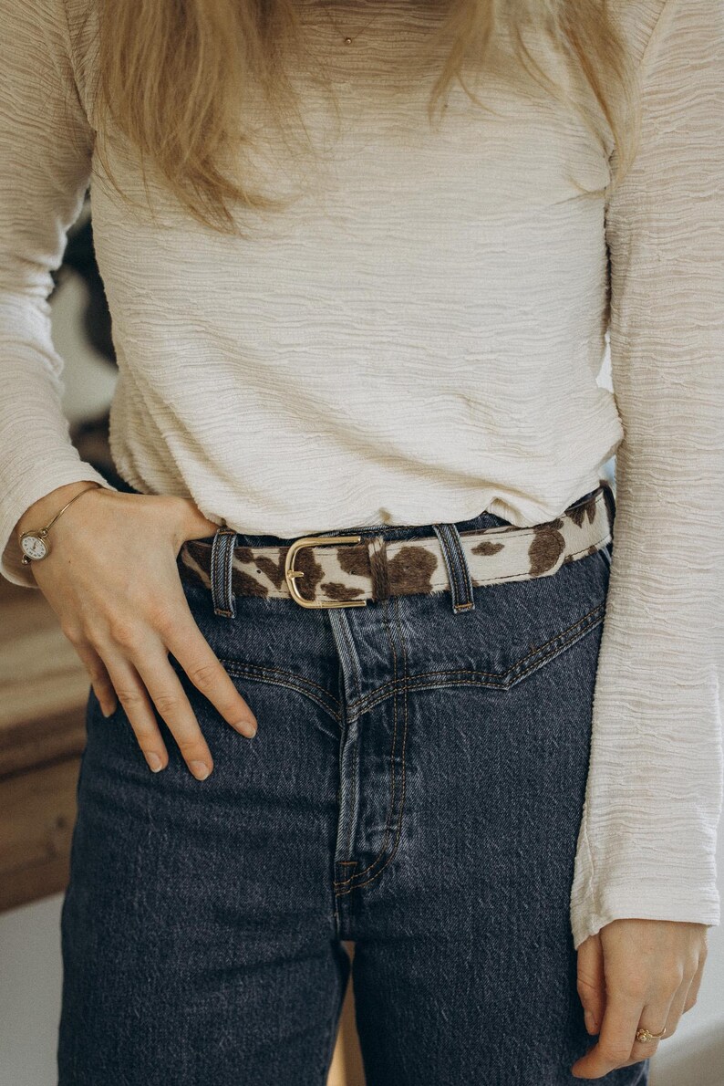 Pode incluir: Um cinto com estampa de pele de vaca e fivela dourada &eacute; usado com jeans azuis e uma camisa branca de manga comprida. O cinto apresenta um padr&atilde;o de manchas marrons e brancas. A fivela tem um design cl&aacute;ssico e arredondado.