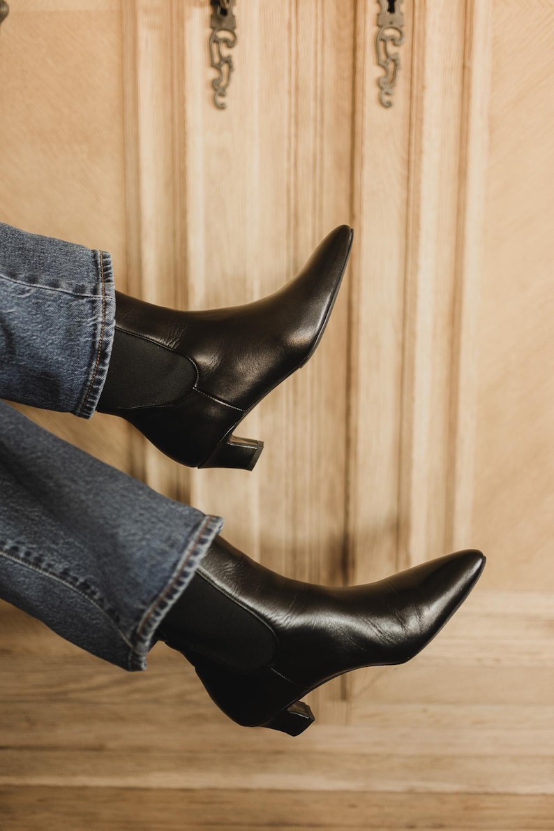Puede incluir: Un par de botines de cuero negro con punta afilada, tac&oacute;n bajo de bloque y paneles laterales el&aacute;sticos, combinados con pantalones vaqueros azules. Las botas presentan un acabado liso y una silueta elegante que llega por encima del tobillo.