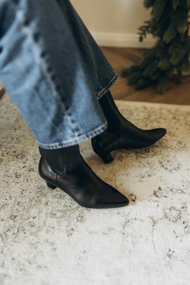 Puede incluir: Un par de botines de cuero negro con punta afilada y tac&oacute;n bajo. Los botines cuentan con paneles el&aacute;sticos laterales y se llevan con pantalones vaqueros azules. La construcci&oacute;n de cuero negro liso ofrece un aspecto limpio y estilizado.