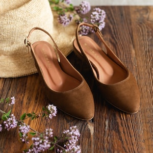 May include: A pair of brown suede slingback flats with a pointed toe and a small buckle on the heel strap. The interior of the shoes features the brand name Munmash printed in gold lettering on the tan insole.
