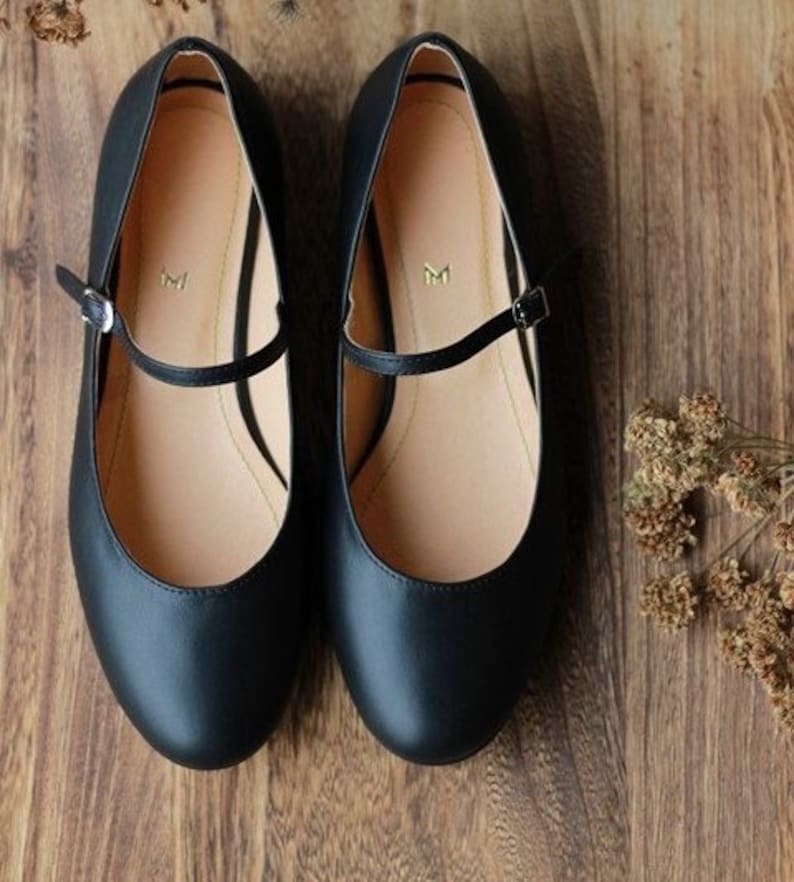 May include: A pair of black leather ballet flats with a strap across the top. The strap has a small gold buckle. The shoes have a rounded toe.