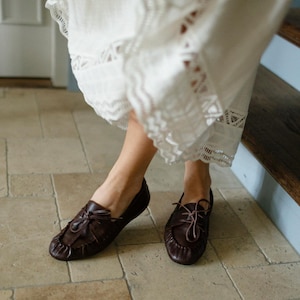 Puede incluir: Un par de mocasines de cuero marr&oacute;n oscuro con dise&ntilde;o de punta fruncida, cordones frontales con tiras finas de cuero y silueta de perfil bajo. Los zapatos se combinan con una falda blanca que presenta un intrincado encaje en el dobladillo.