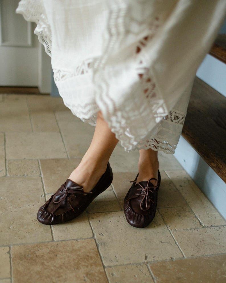 Puede incluir: Un par de mocasines de cuero marr&oacute;n oscuro con dise&ntilde;o de punta fruncida, cordones frontales con tiras finas de cuero y silueta de perfil bajo. Los zapatos se combinan con una falda blanca que presenta un intrincado encaje en el dobladillo.