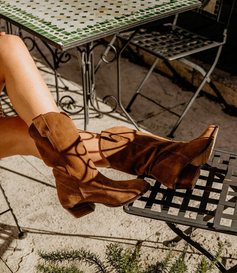 K&ouml;nnte beinhalten: Ein Paar braune Wildleder-Cowgirl-Stiefel mit spitzer Zehenpartie und niedrigem Absatz. Die Stiefel werden von einer Person getragen, die auf einem Metallstuhl mit einem gekachelten Tisch im Hintergrund sitzt.