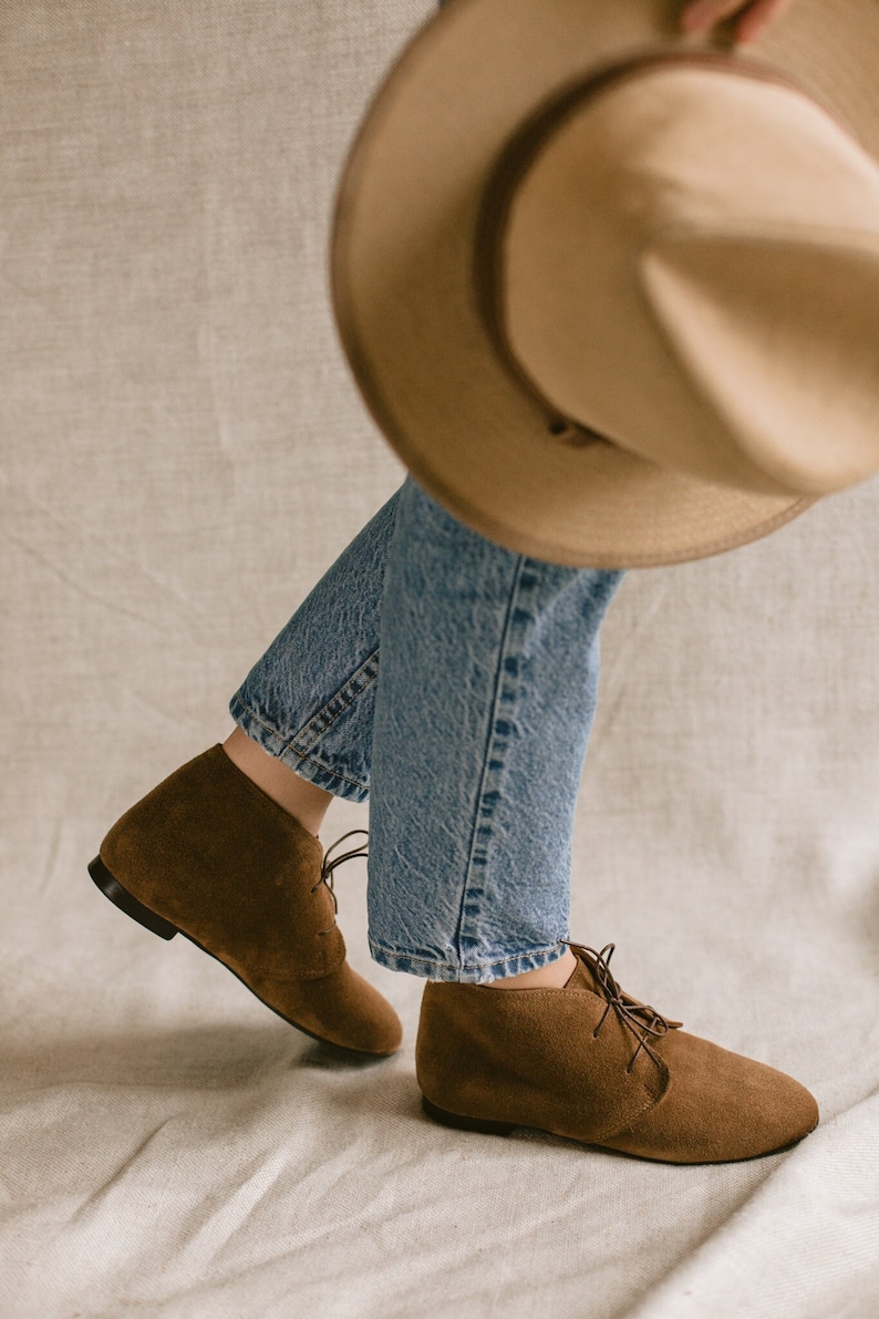May include: A pair of brown suede ankle boots with laces. The boots are worn by a person wearing blue jeans and a brown hat.
