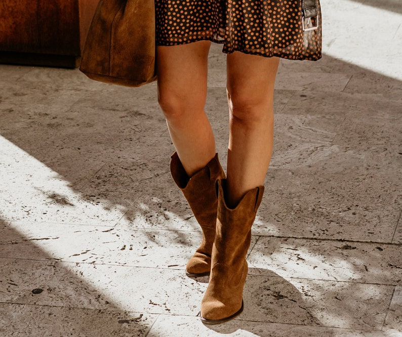 K&ouml;nnte beinhalten: Braune Wildleder-Cowgirl-Stiefel mit spitzer Zehenpartie und niedrigem Absatz. Die Stiefel werden von einer Person getragen, die auf einem Steinboden steht.
