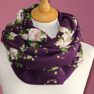 Puede incluir: Una bufanda infinita morada con estampado floral con rosas blancas y hojas verdes.