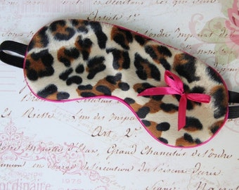 Può includere: Una mascherina per dormire con motivo leopardato marrone, nero e beige su una superficie dalla consistenza morbida. La mascherina è rifinita con un bordo rosa acceso e presenta un piccolo fiocco in nastro rosa abbinato sul lato destro, fissato a un elastico nero.
