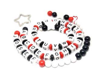 Könnte beinhalten: Eine weiße und schwarze Alphabet-Perlenkette mit dazwischenliegenden roten Perlen. Die Kette hat einen silbernen sternförmigen Anhänger.