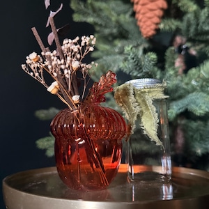 Puede incluir: Un florero de vidrio naranja translúcido con forma de bellota que contiene flores secas y ramitas, junto a un frasco de vidrio transparente con tapa de metal plateado y una cinta de terciopelo verde oliva deshilachada.