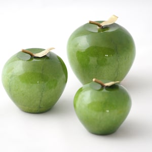 May include: Three green ceramic apples of varying sizes with gold leaf stems. The apples are glossy and have a smooth surface.