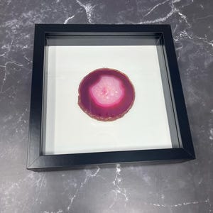 May include: A framed, polished agate slice. The agate features a vibrant pink and white center, surrounded by a deep magenta border. The stone is set against a white background within a black frame, creating a striking contrast. The frame is rectangular.