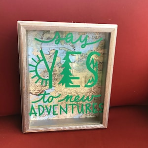 May include: A framed wall art piece with a vintage map background. The green text reads "Say YES to new ADVENTURES" with a sun and tree graphic. The frame is a light, weathered wood.