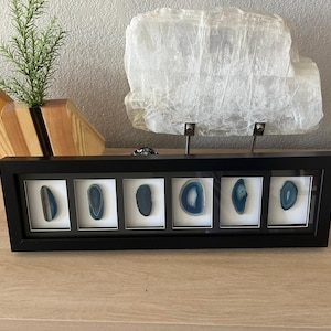 May include: A black framed display featuring six blue and white agate slices. The agate slices are oval-shaped and set against a white background. A large, translucent crystal and a wooden vase with greenery are in the background.