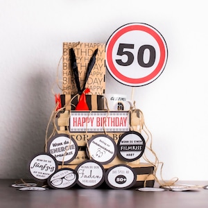 May include: A gift basket for a 50th birthday with a red and white speed limit sign with the number 50, a brown and black gift bag with the words "Happy Birthday" printed on it, a mug with the words "Old Time" printed on it, and several round black and white gift tags with humorous sayings.