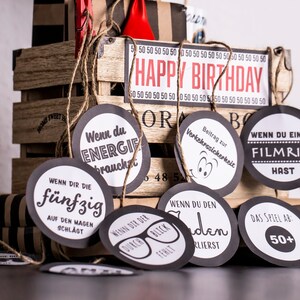 May include: A set of black and white circular gift tags with various phrases in German. The tags are tied together with twine and are hanging from a wooden crate with the words "Happy Birthday" on the front.