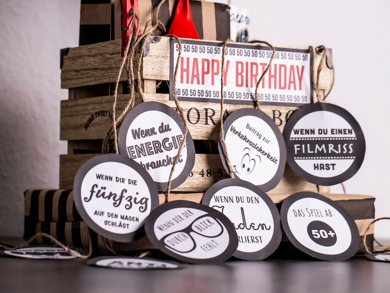 May include: A set of black and white circular gift tags with various phrases in German. The tags are tied together with twine and are hanging from a wooden crate with the words "Happy Birthday" on the front.