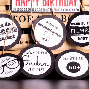 May include: Black and white circular game cards with various phrases and images. The cards are strung on twine and hang from a wooden crate. The phrases on the cards include "When you need energy", "When you lose the thread", "When you have a film tear", "Contribution to road safety", and "The game is over: 50+".