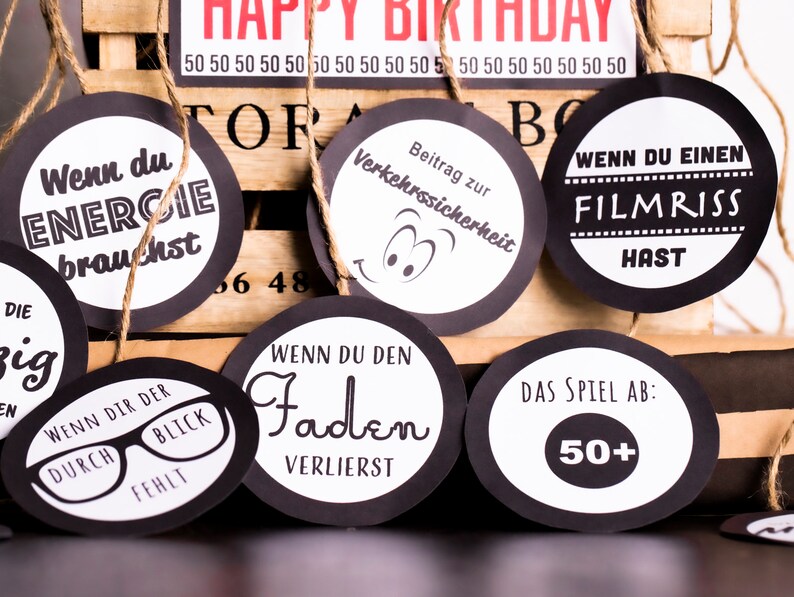 May include: Black and white circular game cards with various phrases and images. The cards are strung on twine and hang from a wooden crate. The phrases on the cards include "When you need energy", "When you lose the thread", "When you have a film tear", "Contribution to road safety", and "The game is over: 50+".