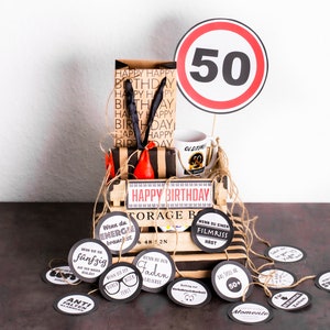 May include: A brown gift box with a black and white striped lid and the words "Happy Birthday" on the front. The box is decorated with a red and black tool, a white mug with the number 50 on it, and a red and white traffic sign with the number 50 on it. The box is surrounded by small round tags with various sayings on them.