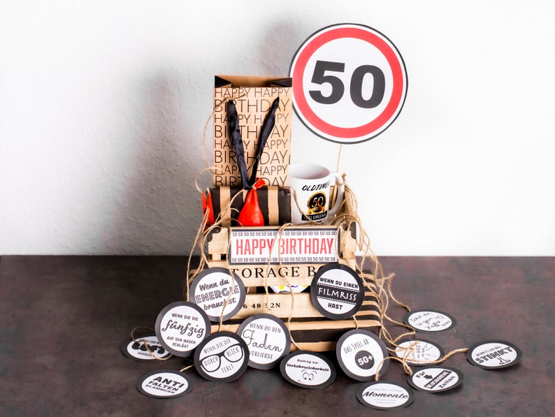 May include: A brown gift box with a black and white striped lid and the words "Happy Birthday" on the front. The box is decorated with a red and black tool, a white mug with the number 50 on it, and a red and white traffic sign with the number 50 on it. The box is surrounded by small round tags with various sayings on them.